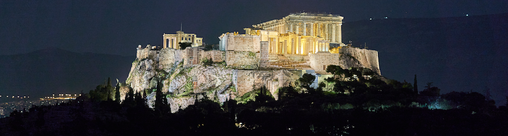 The Acropolis of Athens is home to the Parthenon, a temple dedicated to Αθήνα: the goddess of wisdom and warfare. Picture of the Acropolis at night from the Pnyx with Hymettus in the background by George E. Koronaios adapted from an original on Wikimedia Commons w.wiki/4c2t.